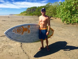 John smiling on the beach with his surfboard.
