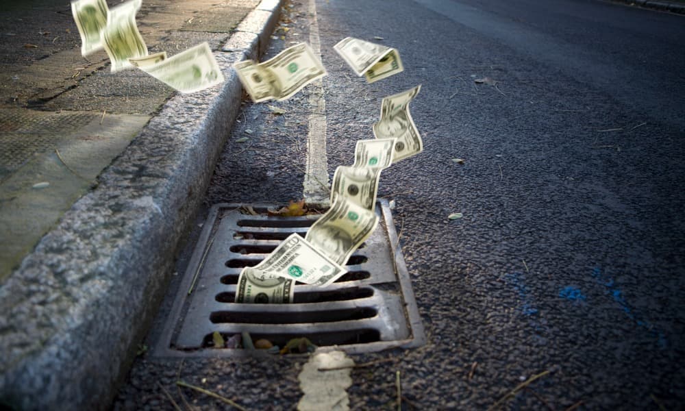 The image displays a street with an up-close view of a gutter. Loads of cash are flowing down from the street and going down the drain. 