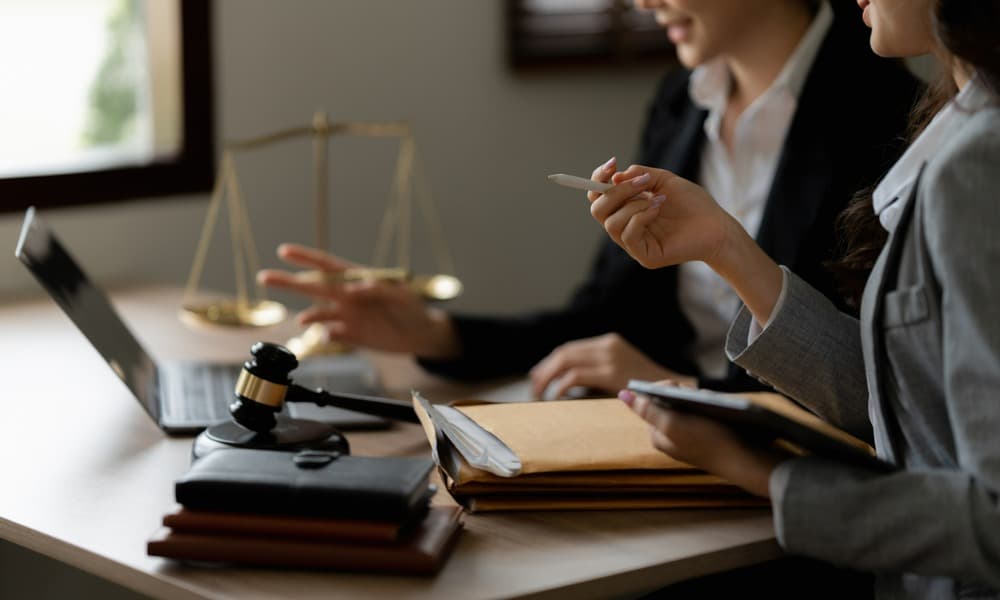 The image shows two female legal professionals working on a laptop atop a desk and discussing documents about a legal case.